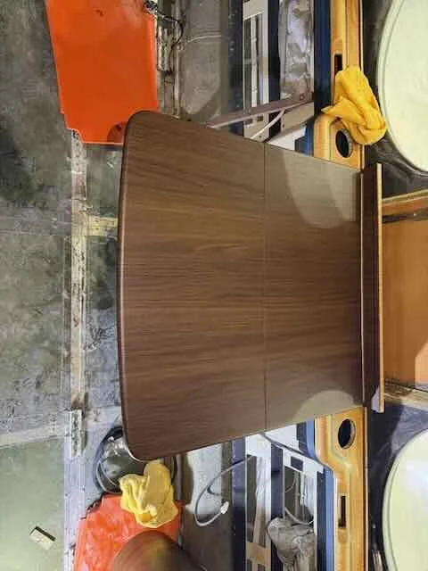 A large wooden table in a workshop or storage area with tools and miscellaneous items nearby.
