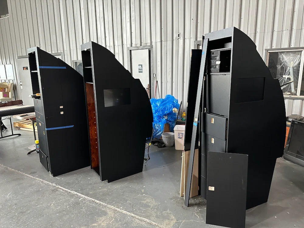 Three black, curved arcade machines in a warehouse or workshop, with some equipment and supplies around.