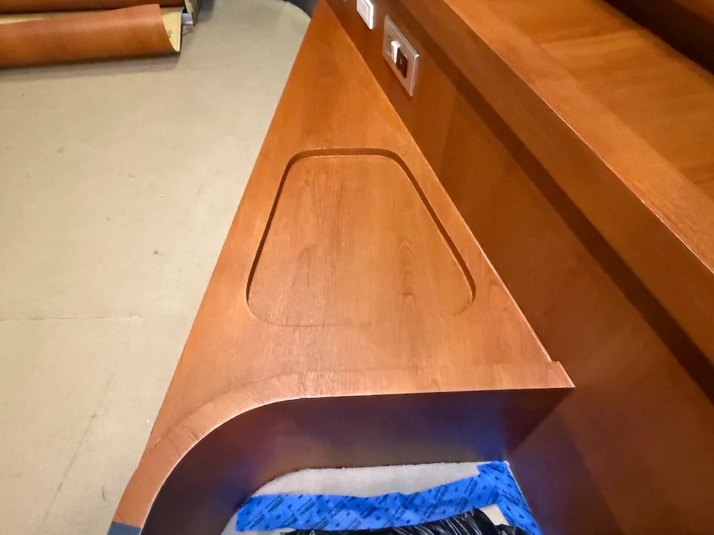 Wooden desk or table with a rectangular cutout and a rounded edge, placed near an electrical outlet on the wall.