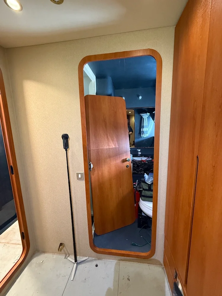 Interior of a room with a wooden door leaning against the wall and a small mirror reflecting part of a cluttered room, including a sink, toiletries, and a towel in the background.
