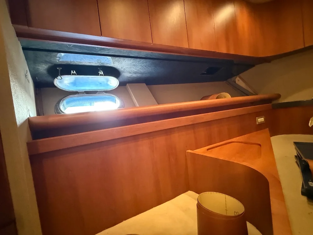 Interior of a yacht with wooden paneling, a small open porthole window letting in natural light, and a seating area with beige cushions.