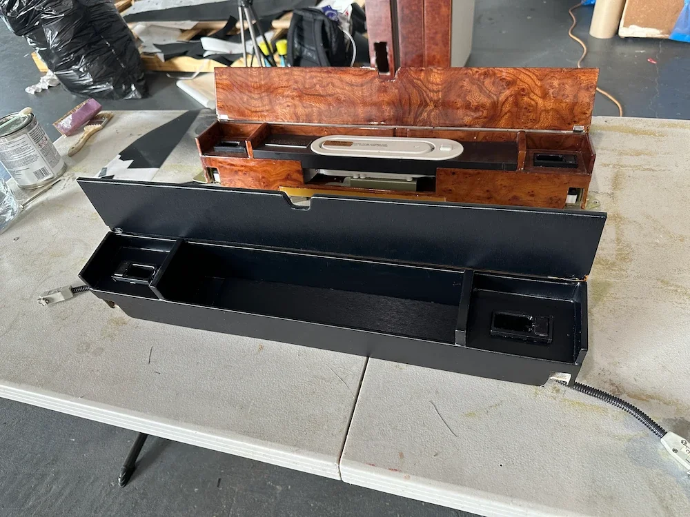 Two vintage car dashboard panels, one with a wood grain finish and the other black, placed on a table in a workshop.