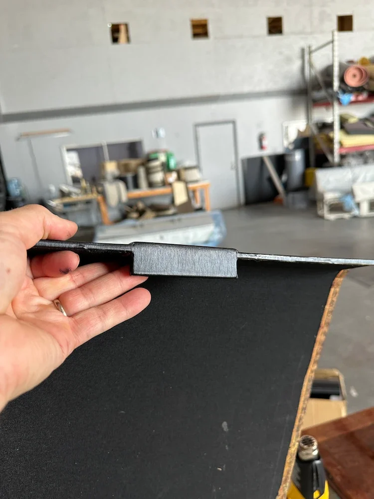 Close-up of a hand holding a black metal object, possibly a shelf or bracket, in front of a workshop with shelves, equipment, and materials in the background.