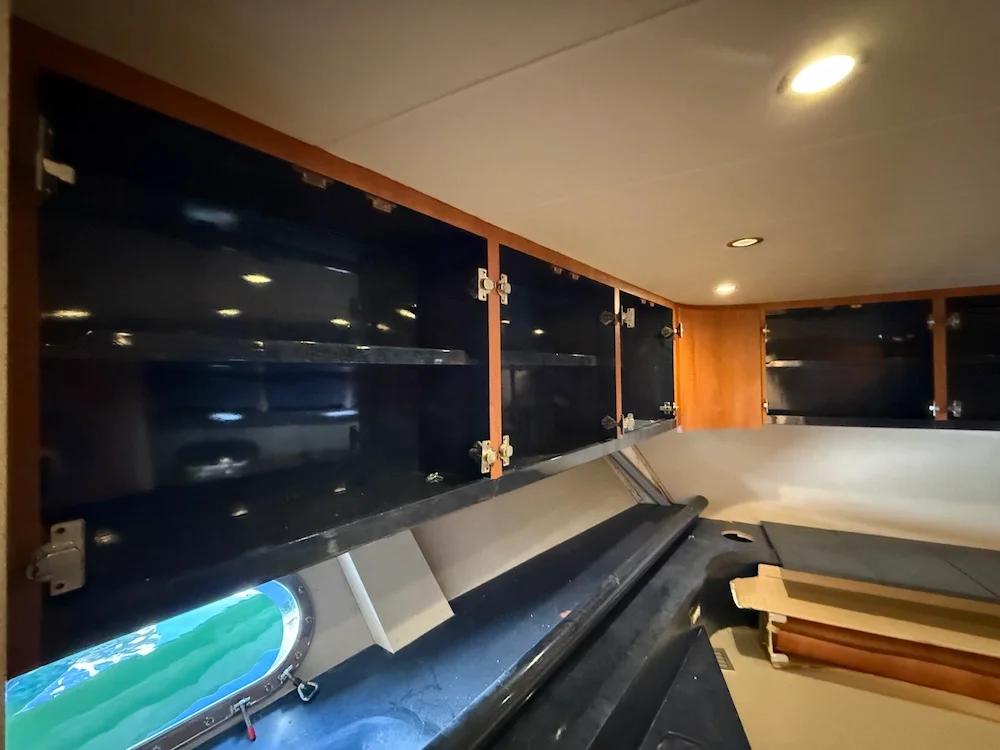 Interior shot of a yacht with empty black cabinets, a window showing water outside, and some stacked wood or material on a black surface.