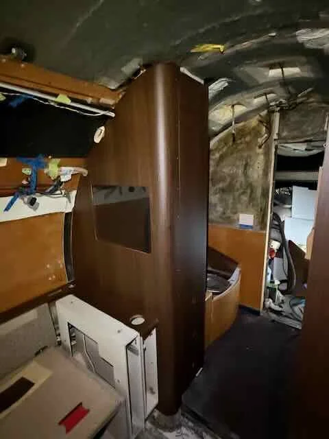 Interior of a small, cluttered space with various appliances and wooden furniture, including a microwave and cabinetry, in a room with a curved ceiling and exposed wiring.