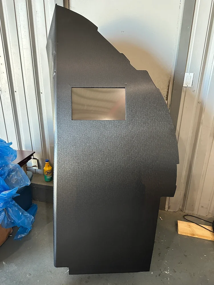 Black arcade machine with a small screen, placed in a warehouse or repair shop with cleaning supplies nearby.