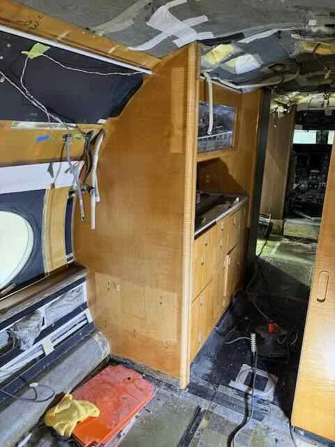 Interior of a camper or RV under renovation with exposed wiring, unfinished surfaces, and a partially installed wooden cabinet and countertop.
