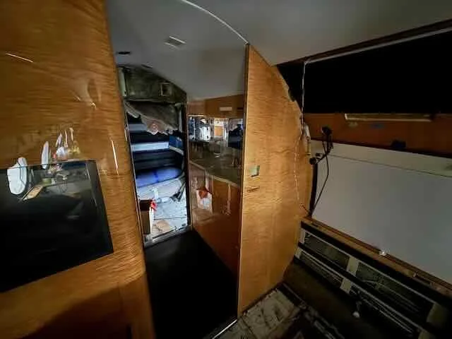 Interior of a small, cluttered RV or camper with a wooden partition, a bed, and a kitchenette with cabinets and a countertop.
