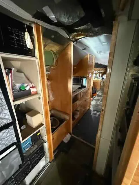 Narrow kitchen with wooden cabinets, microwave, and shelves filled with various items, leading into an RV or caravan interior.