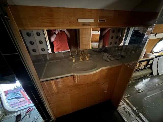 Reflected image of a person in a mirror above a bathroom sink with gold fixtures, wooden cabinets, and a partially visible washer or dryer to the right.