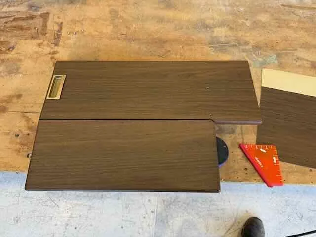 Two wooden laminate flooring planks, a red carpenter's square, and a piece of beige sheet music or paper on a workbench.