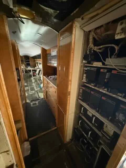 A narrow hallway inside a mobile home or RV with wooden cabinets on the right and a black carpet runner on the floor leading to a room with a small dining area in the background.