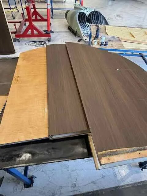 Three wooden table tops in different shades of brown and one yellow are placed on a work table in a woodworking shop. In the background, there are tools, a fan, and other woodworking equipment.