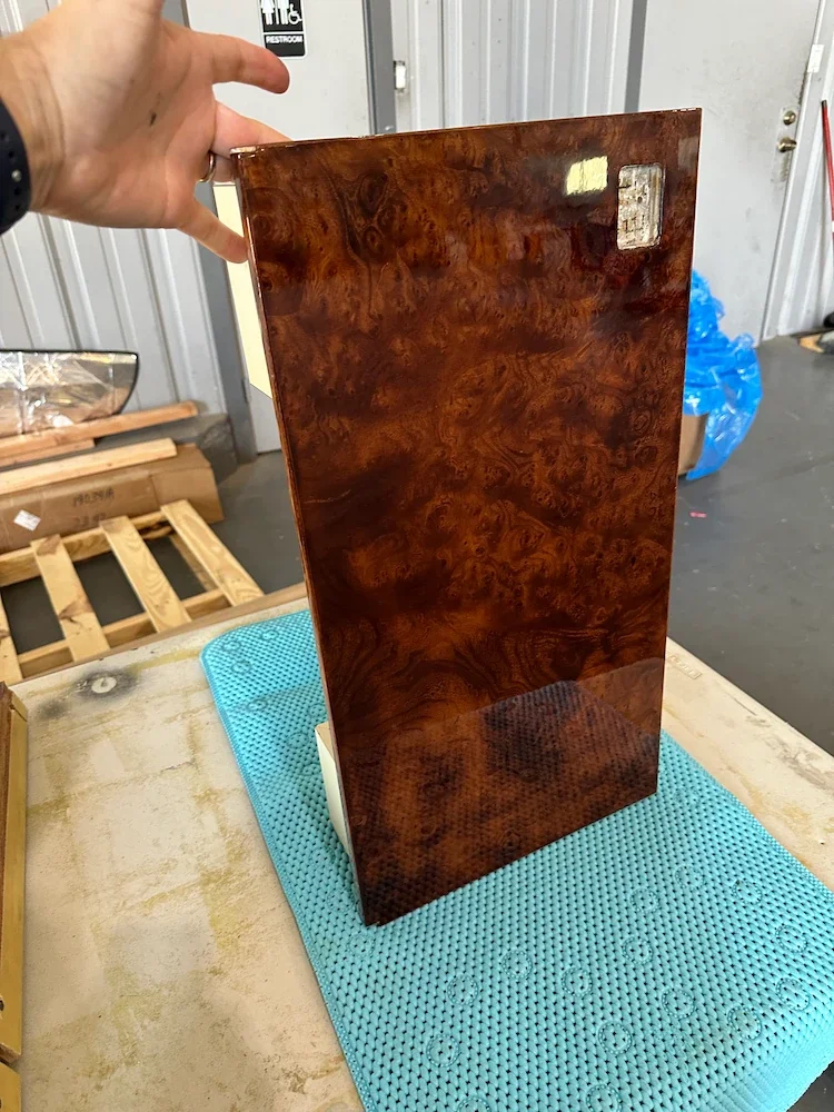 A hand holding a rectangular piece of glossy wood with a burl grain pattern, standing upright on a textured blue surface, in a workshop or storage area.