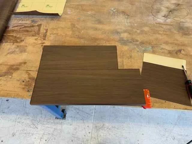 A wooden workbench with several brown wood laminate sheets on it, a yellow sticky note, and a black pen.
