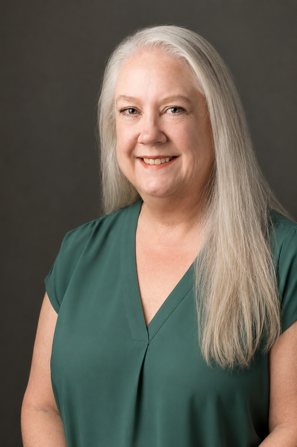 A woman with long gray hair wearing a teal blouse, smiling against a neutral gray background.