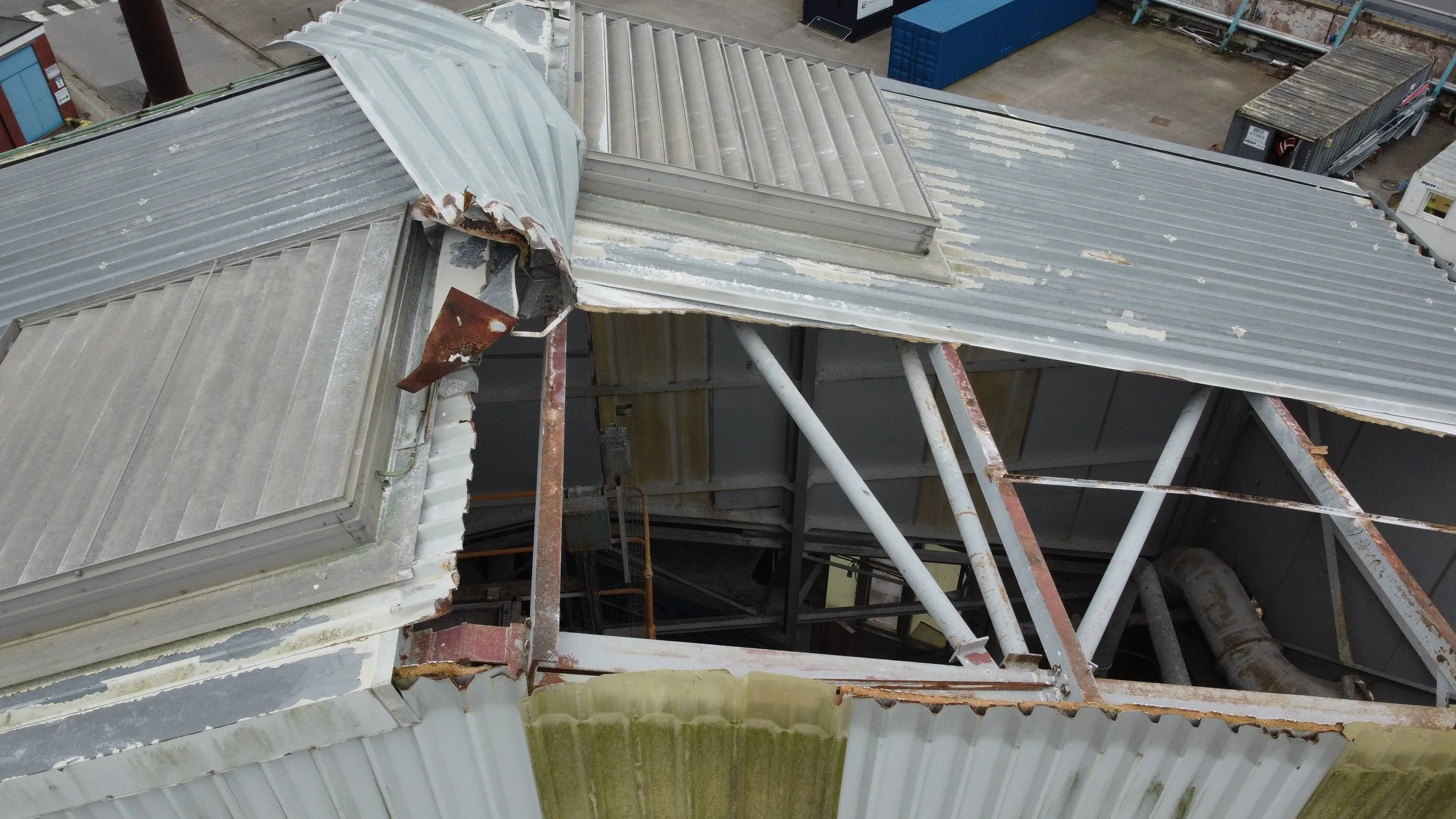 Overhead view of a rooftop with metal and corrugated panels, some of which are damaged or bent, supported by scaffolding. In the background, there are storage containers and construction materials.