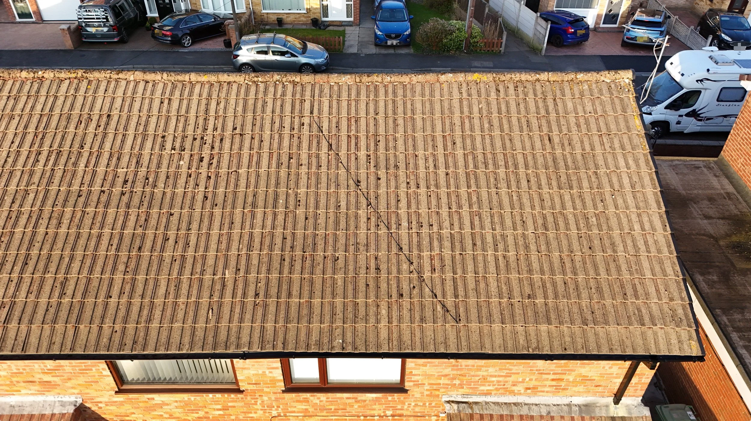 Aerial view of a residential rooftop with visible tiles, neighboring houses, parked cars, and a street with greenery.