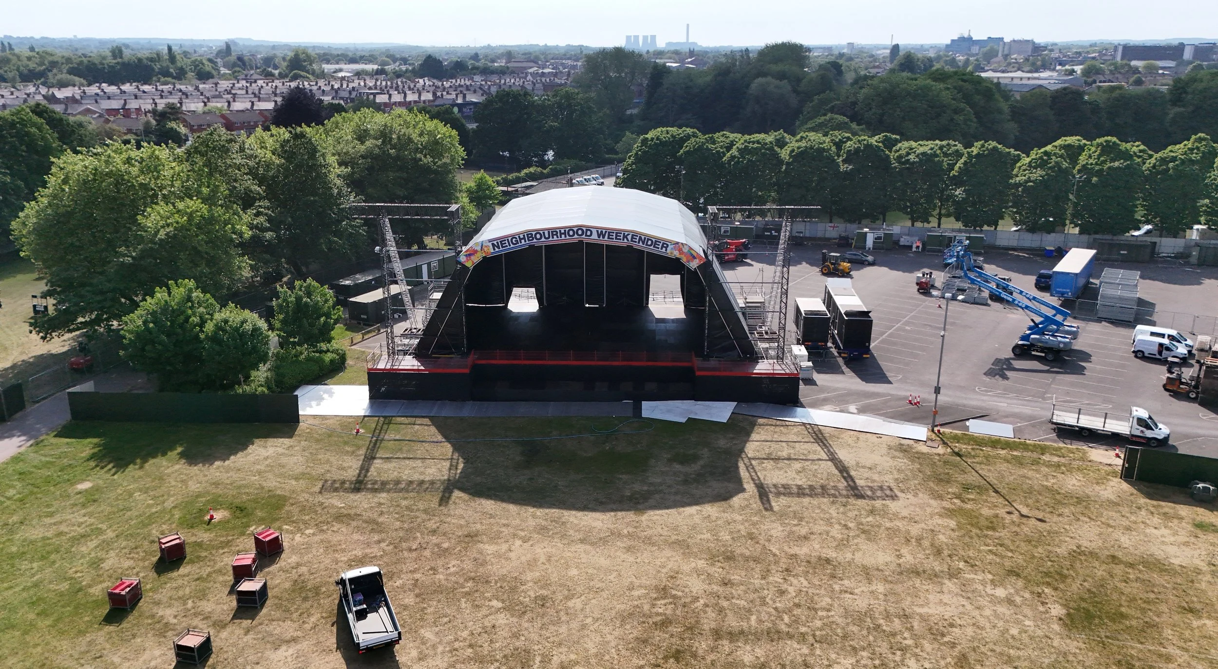 An outdoor stage setup at a community event called 'Neighbourhood Weekender,' with a white canopy, surrounded by trees and parking area, with various trucks and equipment nearby.