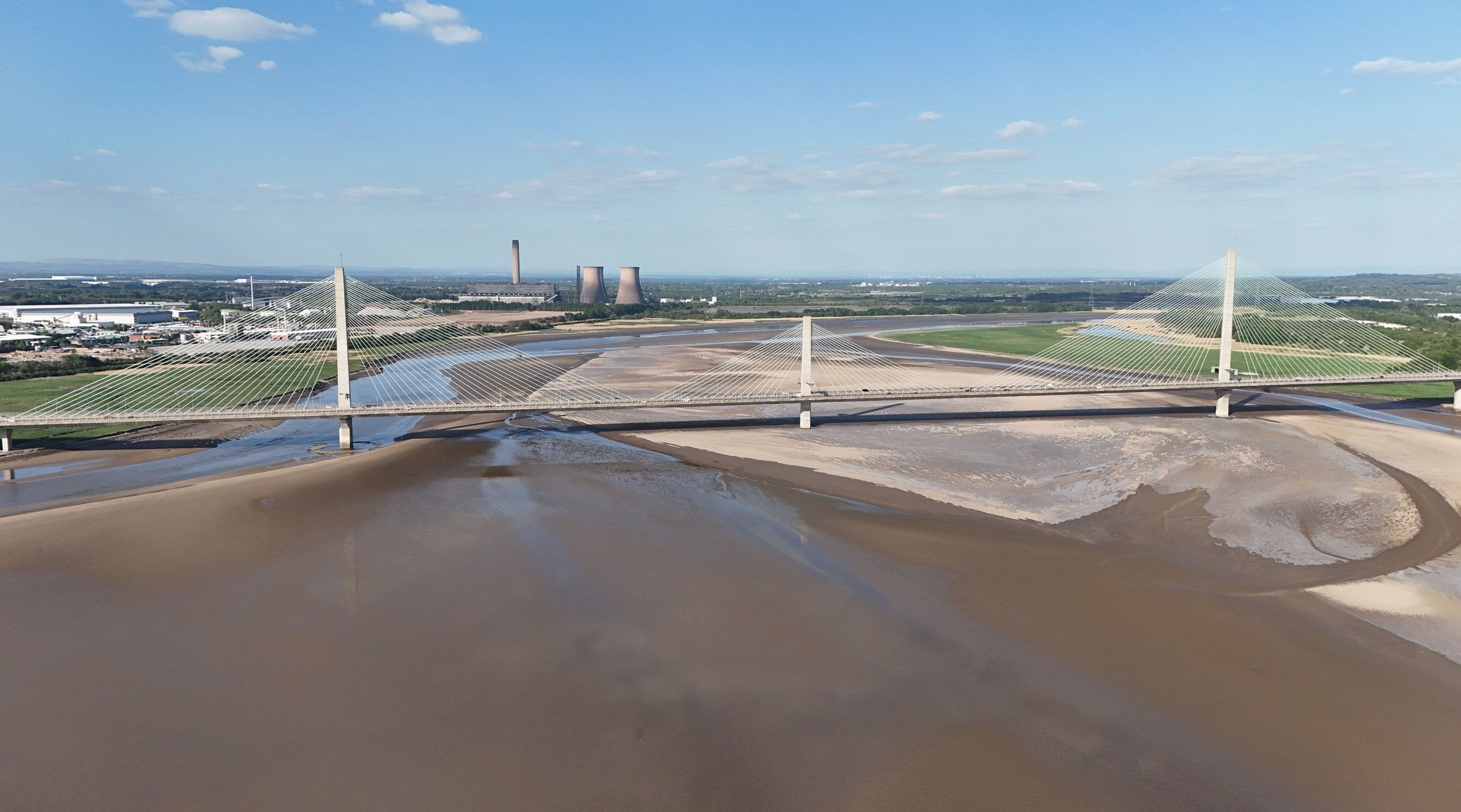 A bridge with cable-stayed design spans over a river or floodplain, with industrial facilities in the background and a partly cloudy sky overhead.