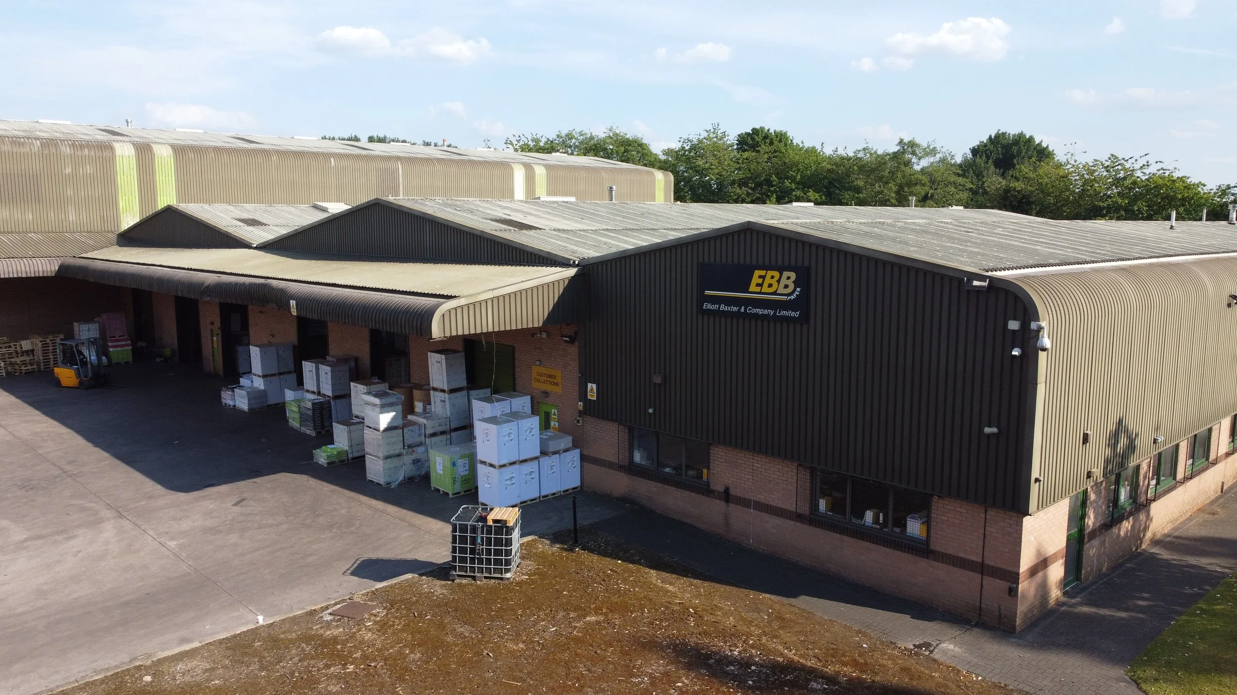 Industrial warehouse building with a sign 'E B B Elliott Baxter & Company Limited'. Exterior includes crates, pallets, and a forklift nearby in an outdoor yard.