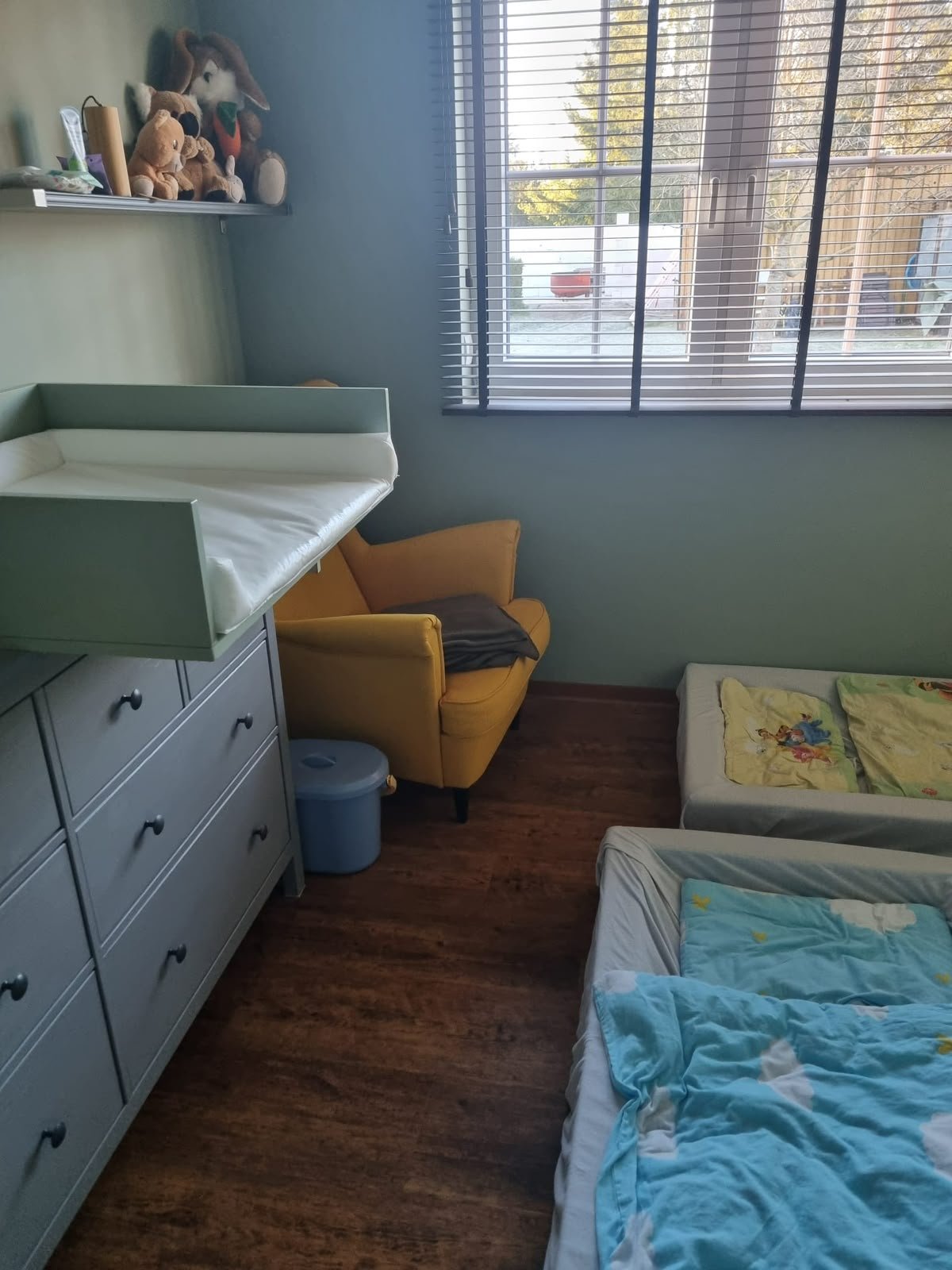 Ein Kinderzimmer mit einem Wickeltisch, einem gelben Sessel, einem Kinderbett mit Disney-Motiv und Fenster mit Jalousien.
