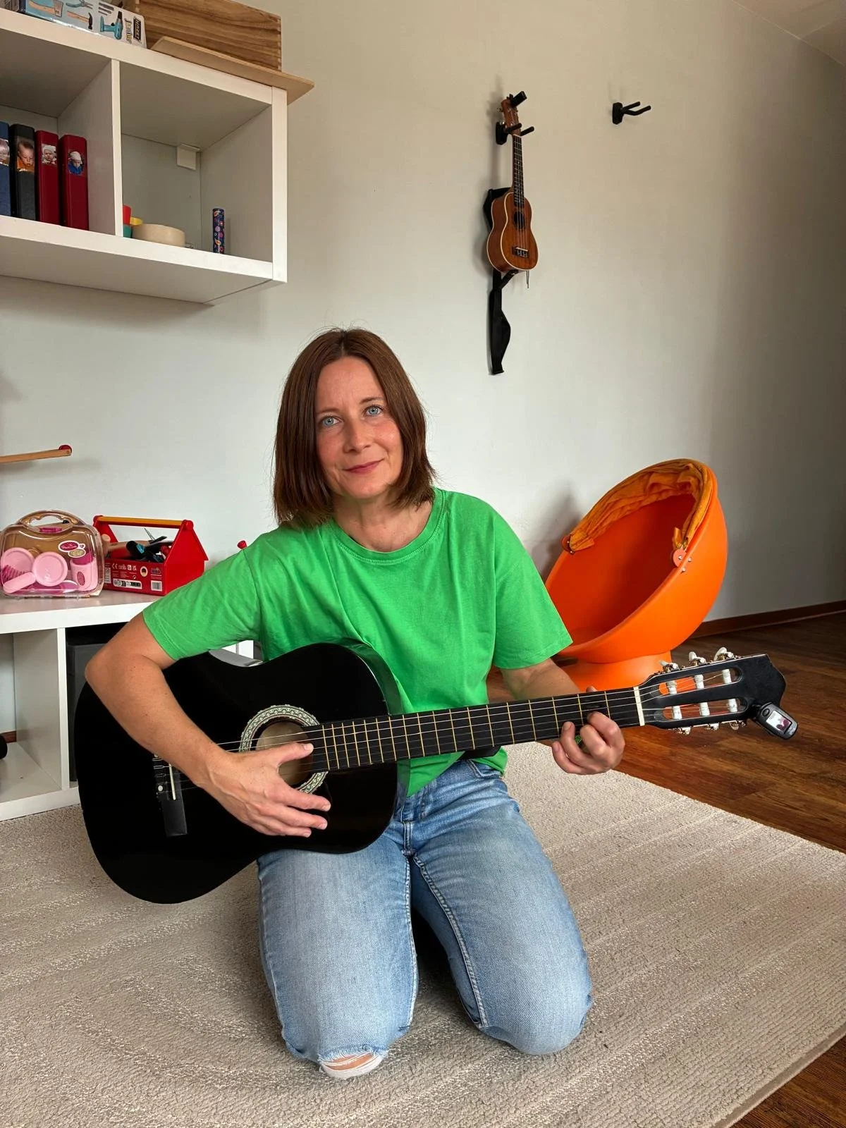 Eine Frau sitzt auf dem Boden, spielt Gitarre und lächelt. Sie trägt ein grünes T-Shirt und Jeans. Im Hintergrund sind ein orangees Sitzmöbel, eine Gitarre an der Wand, und ein Regal mit Spielzeug und Büchern sichtbar.