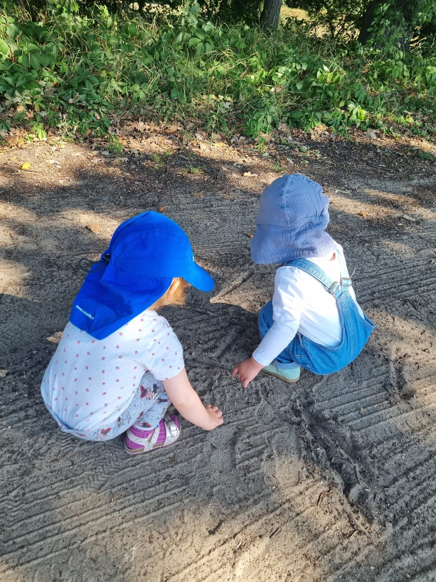 Zwei kleine Kinder knien auf dem Boden im Freien, schauen aufmerksam auf den Boden, während sie im Schatten eines Baumes spielen.