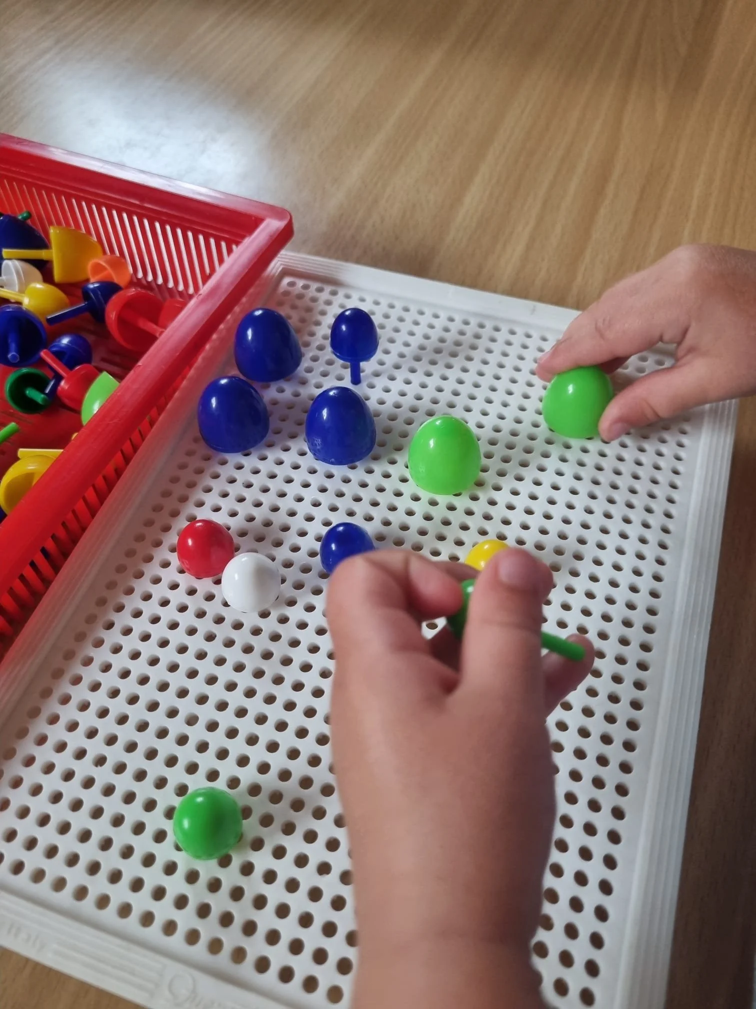 Zwei Hände spielen ein Brettspiel mit bunten Eier- und Pistolenspielfiguren auf einem weißen Spielfeld, daneben ein roter Spielzeugkorb mit weiteren bunten Figuren auf einem Holztisch.