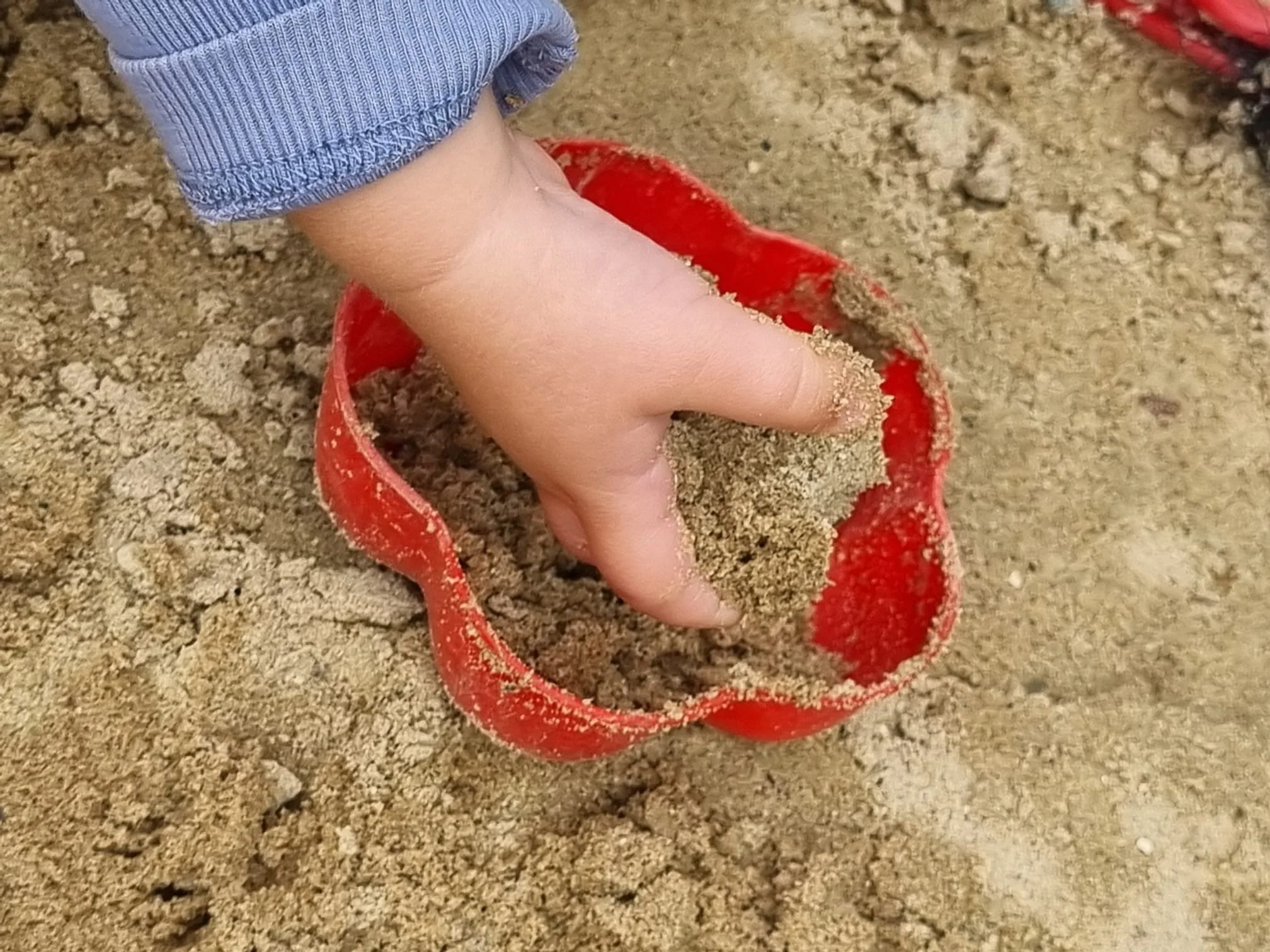 Ein Arm eines Kindes in einem blauen gestreiften Pullover, das mit der Hand in einem roten Sandkasten spielt. Die Hand ist in den Sand getaucht, auch Sand klebt an den Fingern. Der Sandkasten befindet sich auf einer Sandfläche.