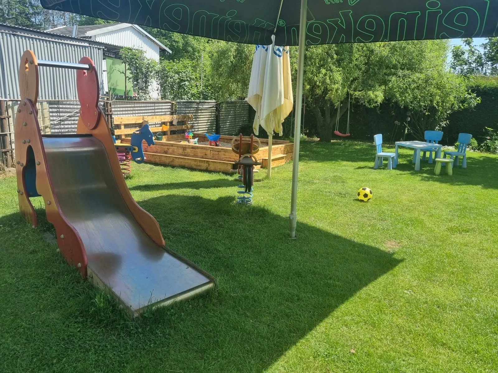 Blick auf einen Kinderspielplatz mit Rutsche, Sandkasten, größeren Bänken und einem Fußball, umgeben von grüner Wiese und Bäumen bei sonnigem Wetter.