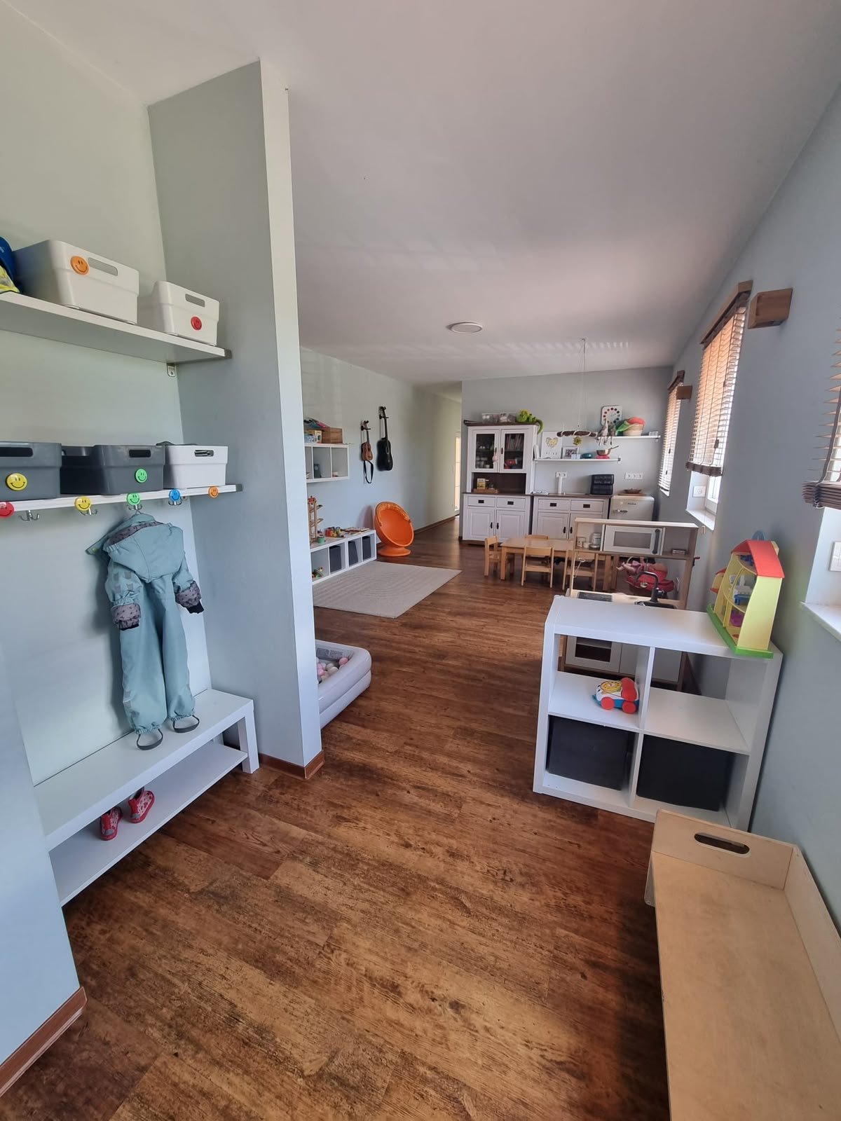 Ein hell erleuchteter Kinderraum mit Holzfußboden, Spielzeugregalen und einem Wickeltisch, mit mehreren Fenstern, die Tageslicht hereinlassen.