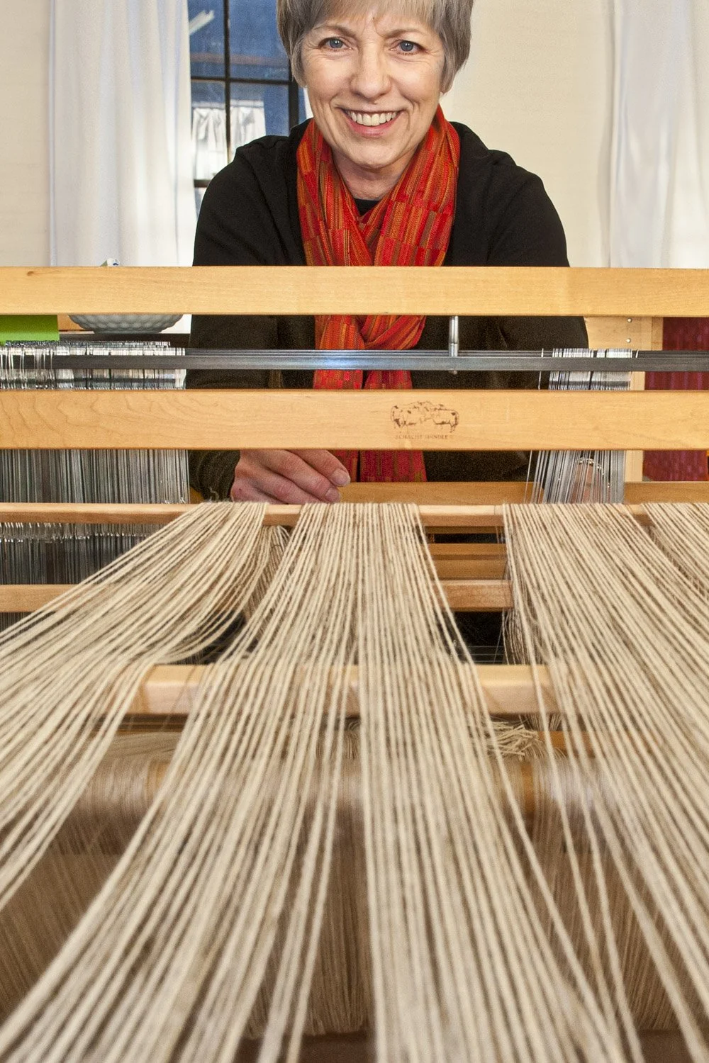 A smiling Karen Donde in her studio, wearing a red scarf and black top sitting behind a loom with multiple rows of beige yarn threads.