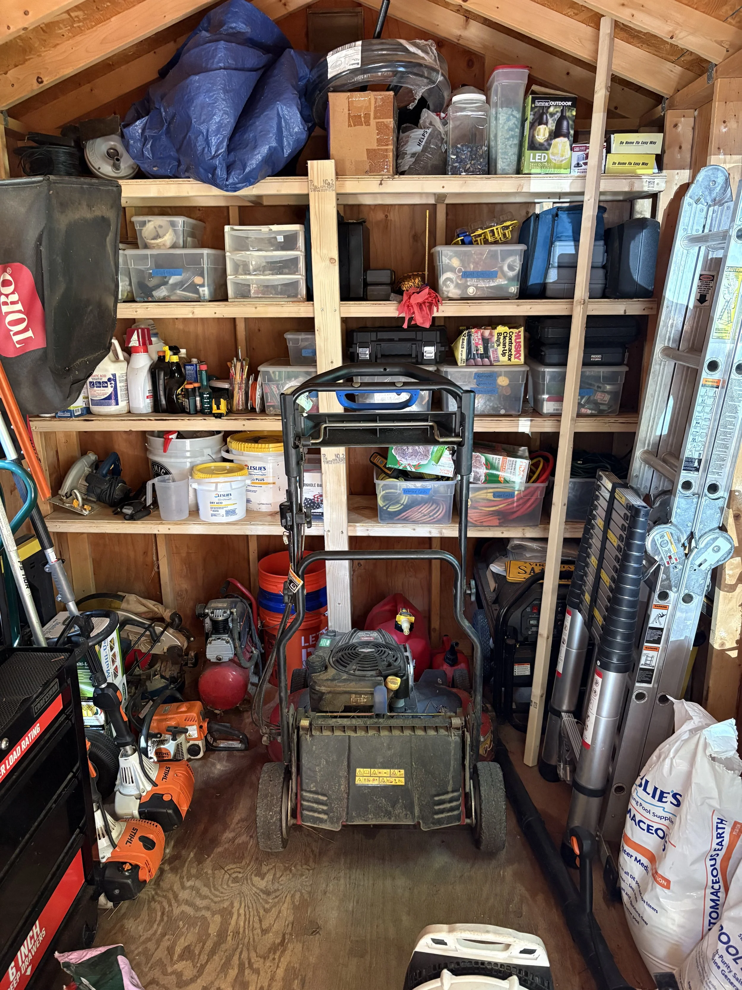 A cluttered storage shed containing shelves filled with tools, supplies, and equipment including a lawnmower, power tools, bags of supplies, and various small containers.