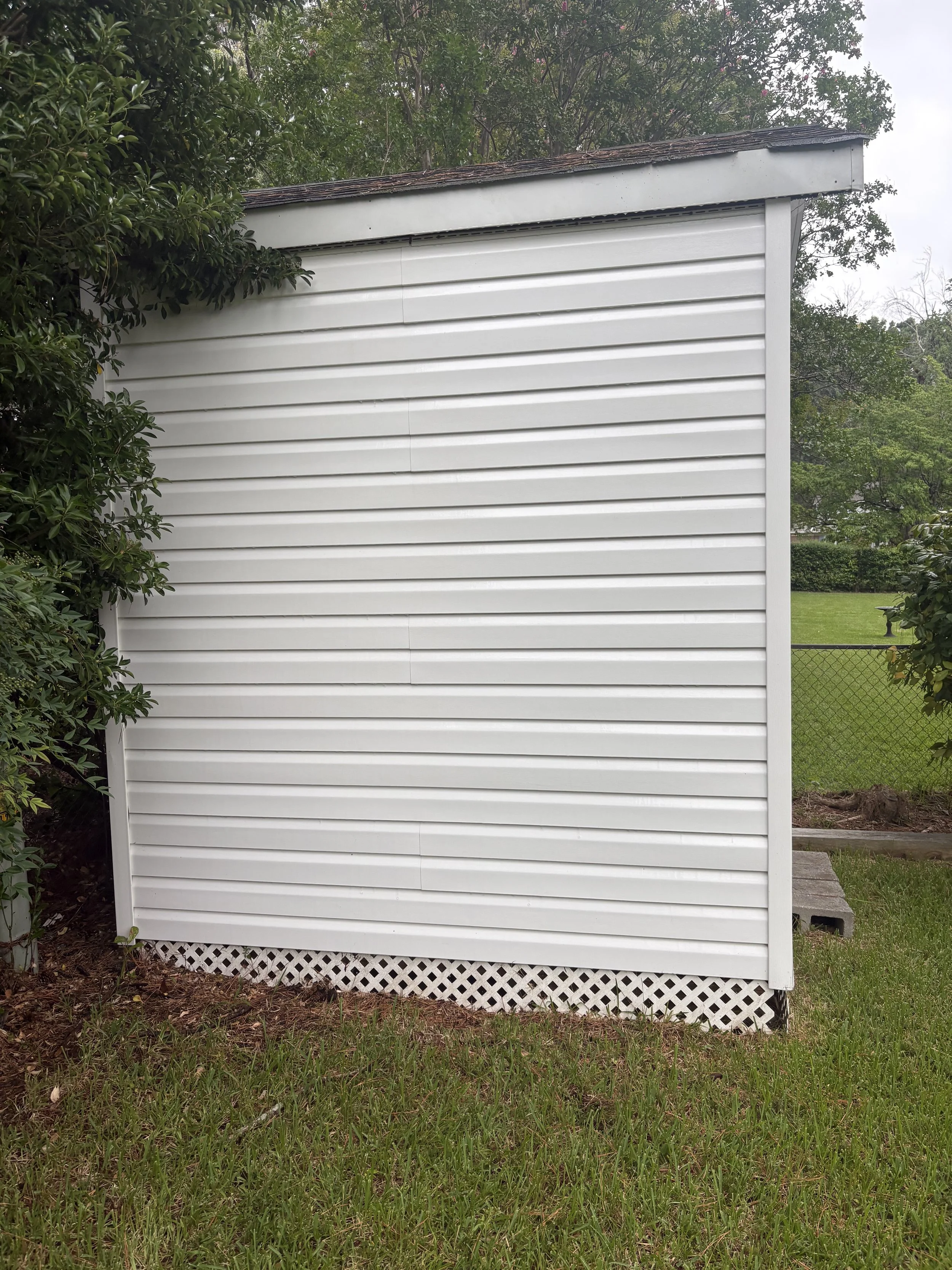 A white shed with horizontal siding, surrounded by greenery and trees in a backyard