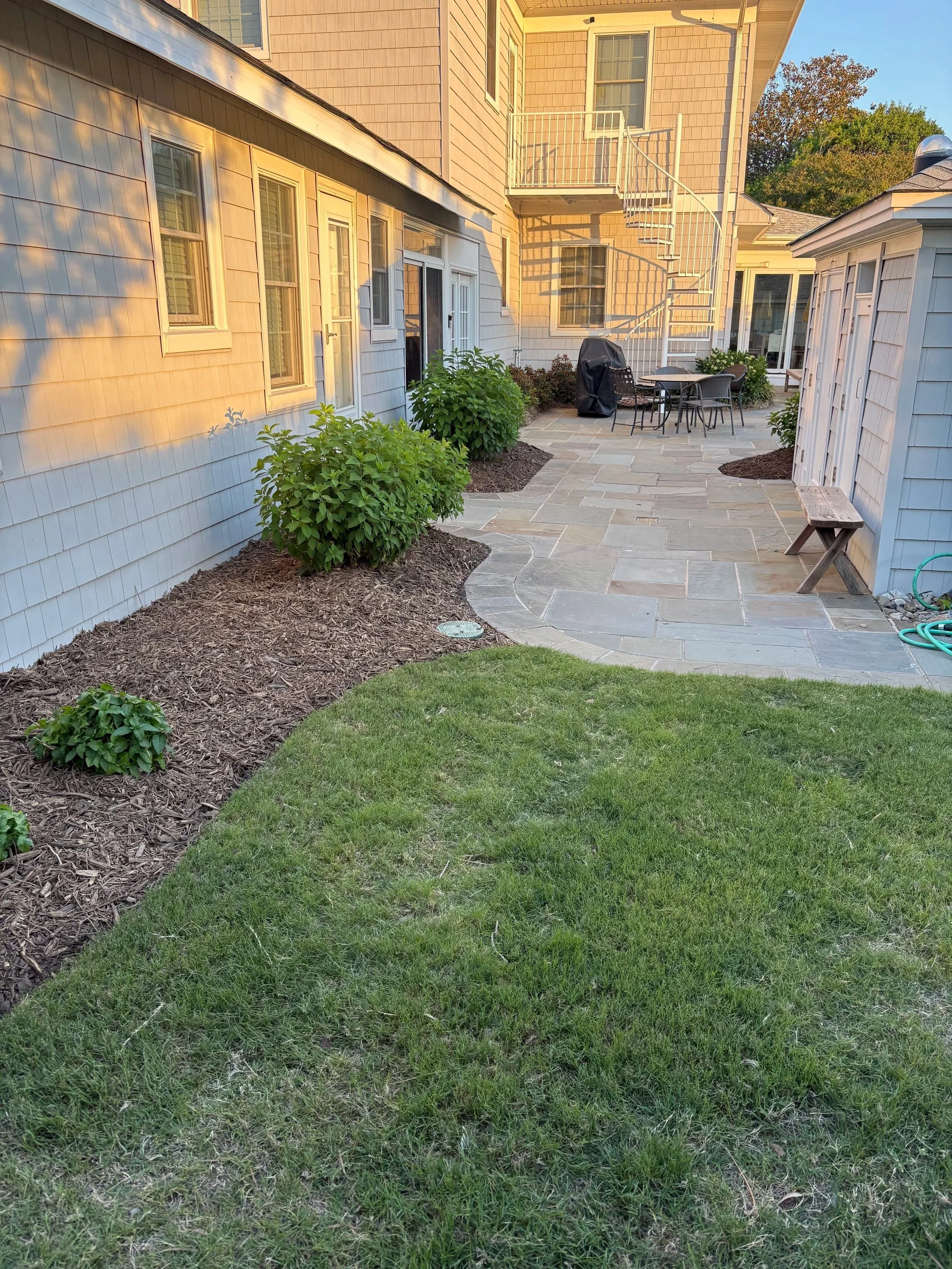 Backyard patio area with a stone pathway, green bushes, a lawn, and a white house with a spiral staircase leading to a second-story balcony.