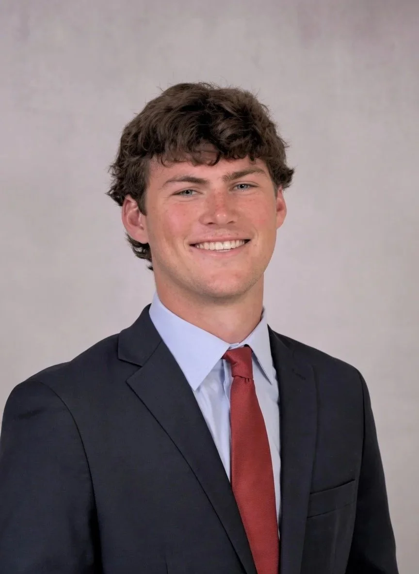 Young man with brown curly hair wearing a dark suit, light blue shirt, and red tie, smiling at the camera.