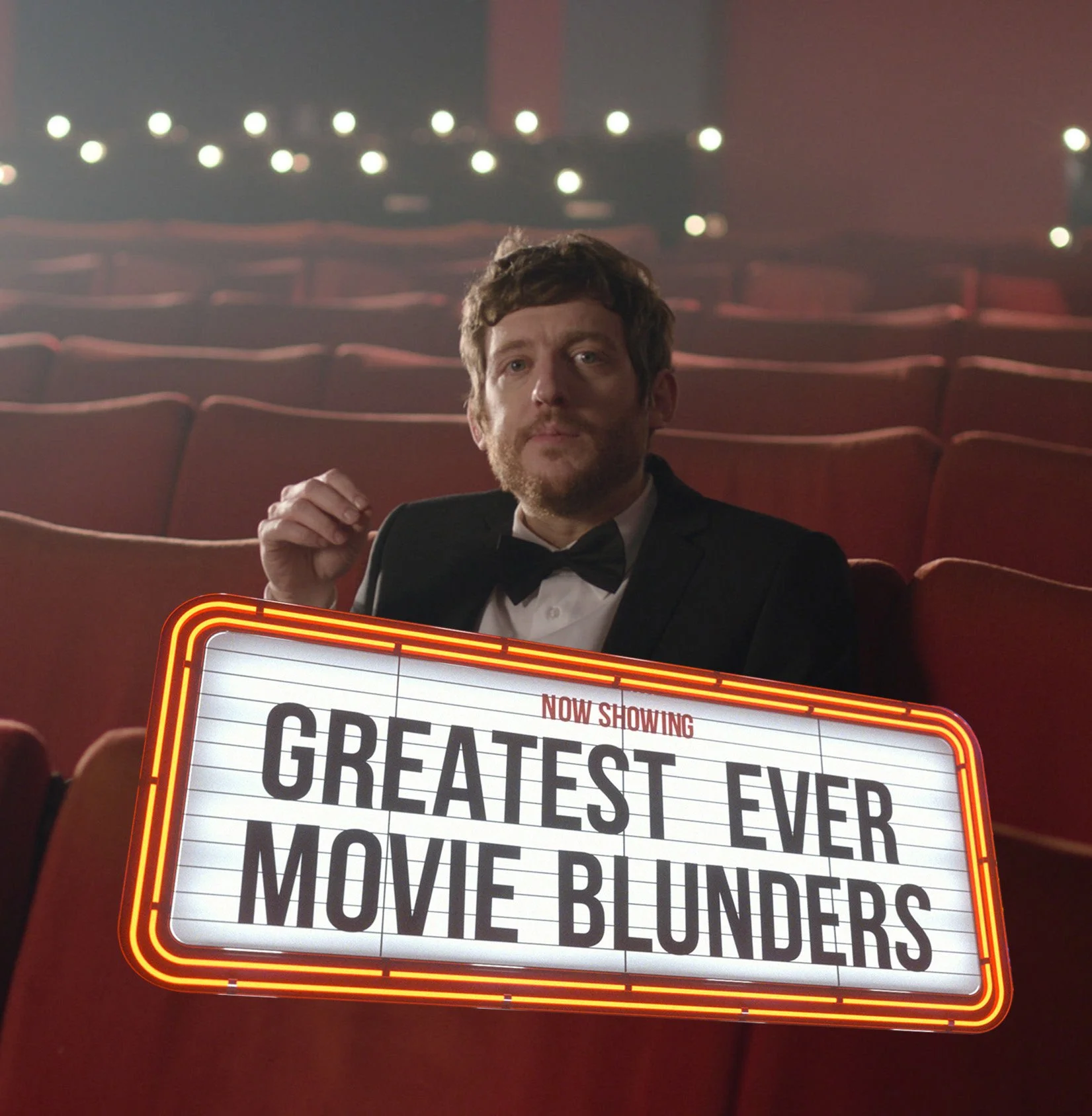 A man in a tuxedo sitting in a theater with red seats holds a marquee sign that reads: 'Now Showing Greatest Ever Movie: Munde Blunders'.