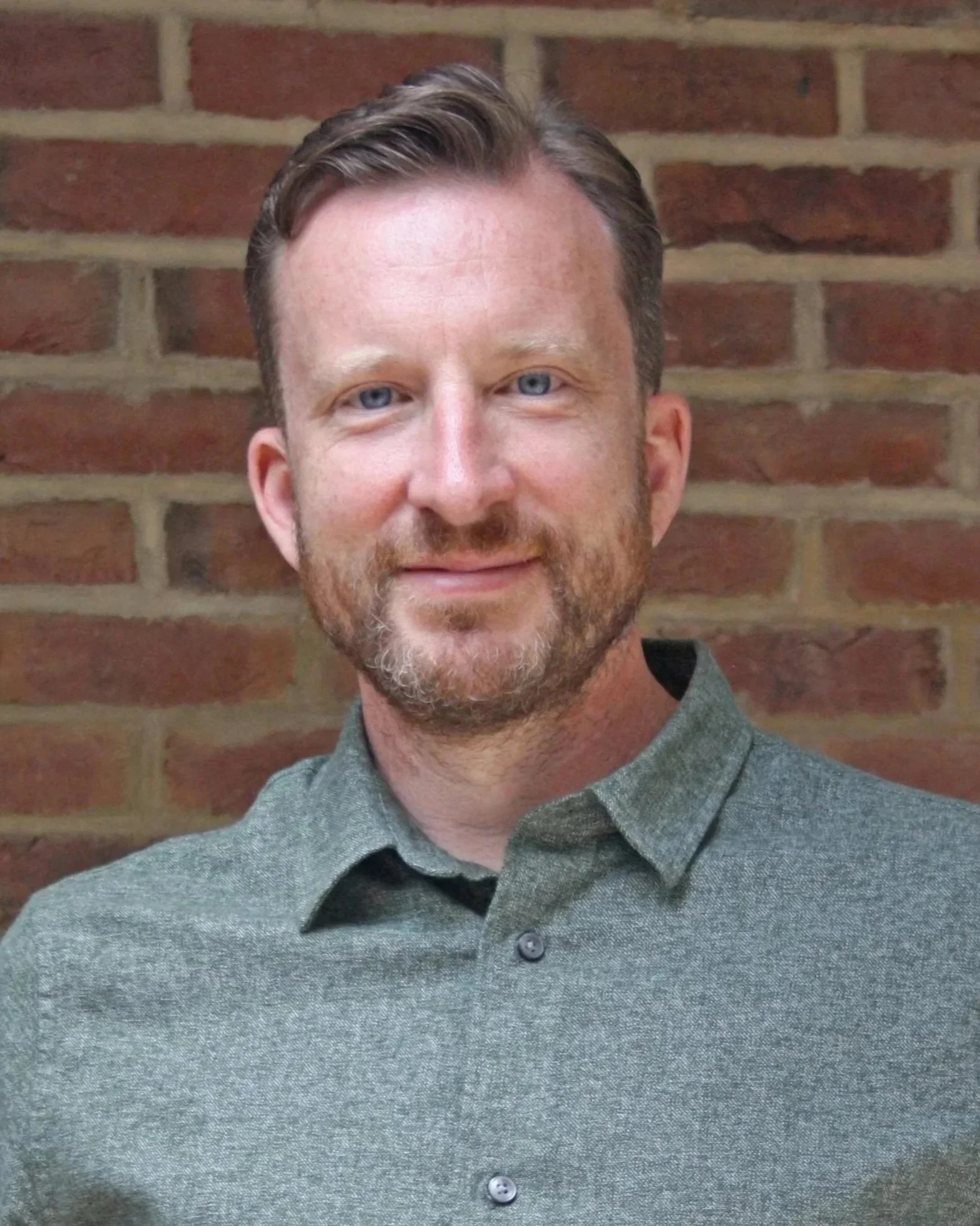 Close-up of a man with blue eyes, light brown hair with streaks of gray, and a beard, wearing a gray shirt, standing against a brick wall background.