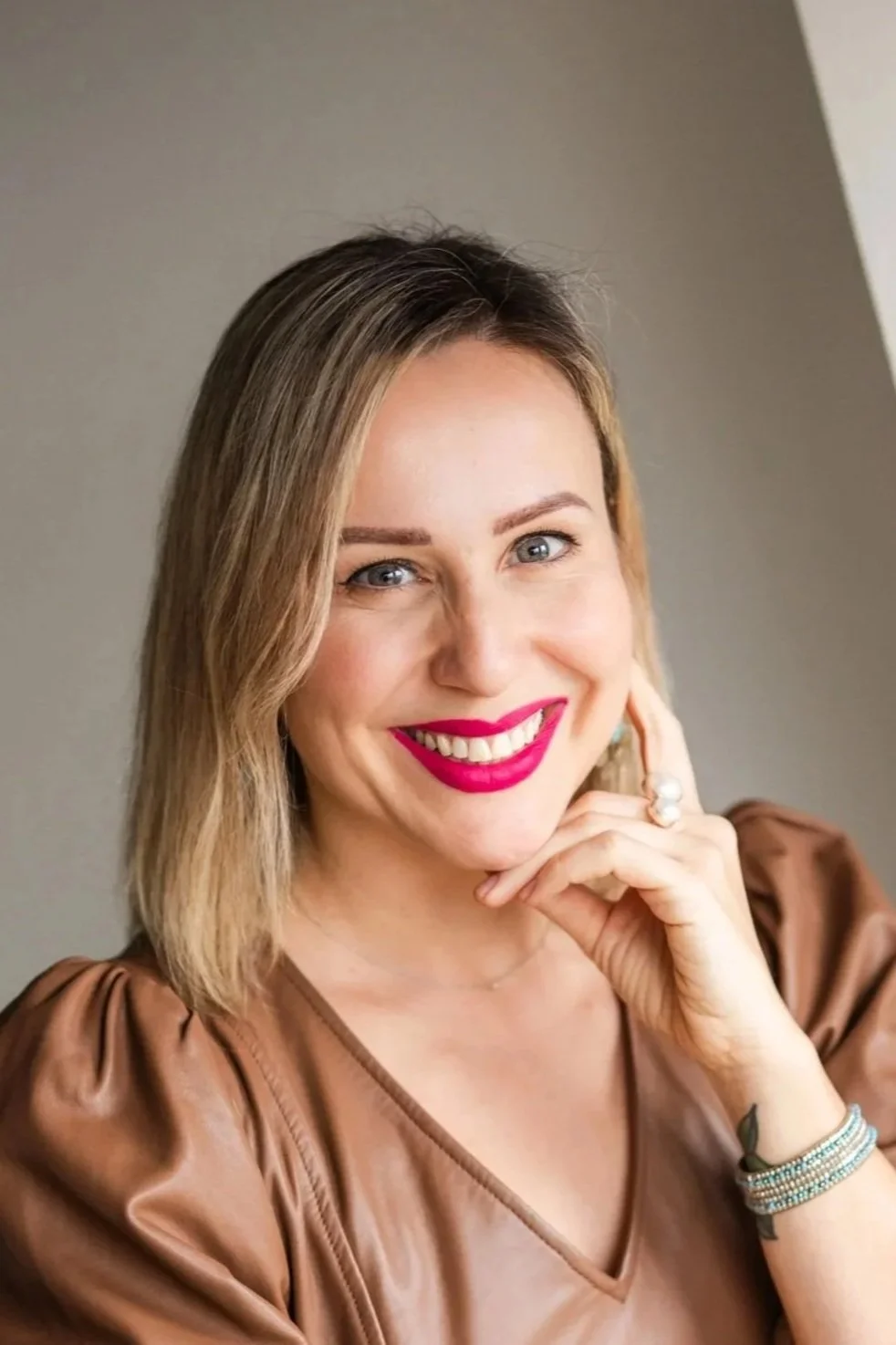 A woman with blonde hair and blue eyes smiling, wearing pink lipstick, a brown top, a ring, and bracelets, leaning her chin on her hand.