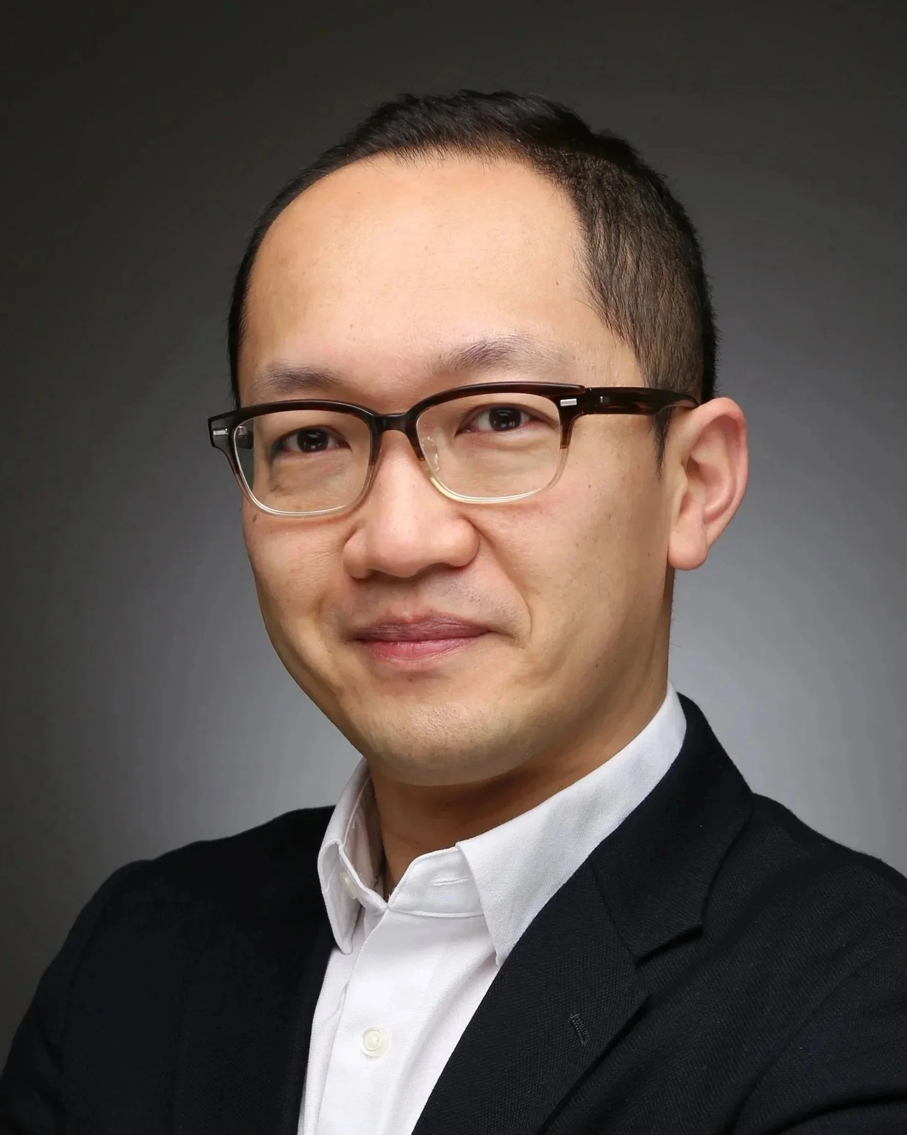 Close-up of a smiling Asian man wearing glasses, a white shirt, and a black sweater, against a dark background.