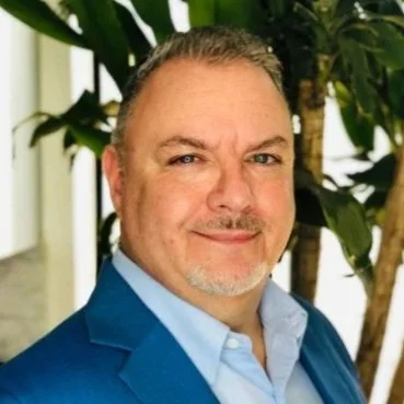 A man with short, dark hair and a goatee, wearing a blue suit jacket and light blue shirt, standing near plants indoors.
