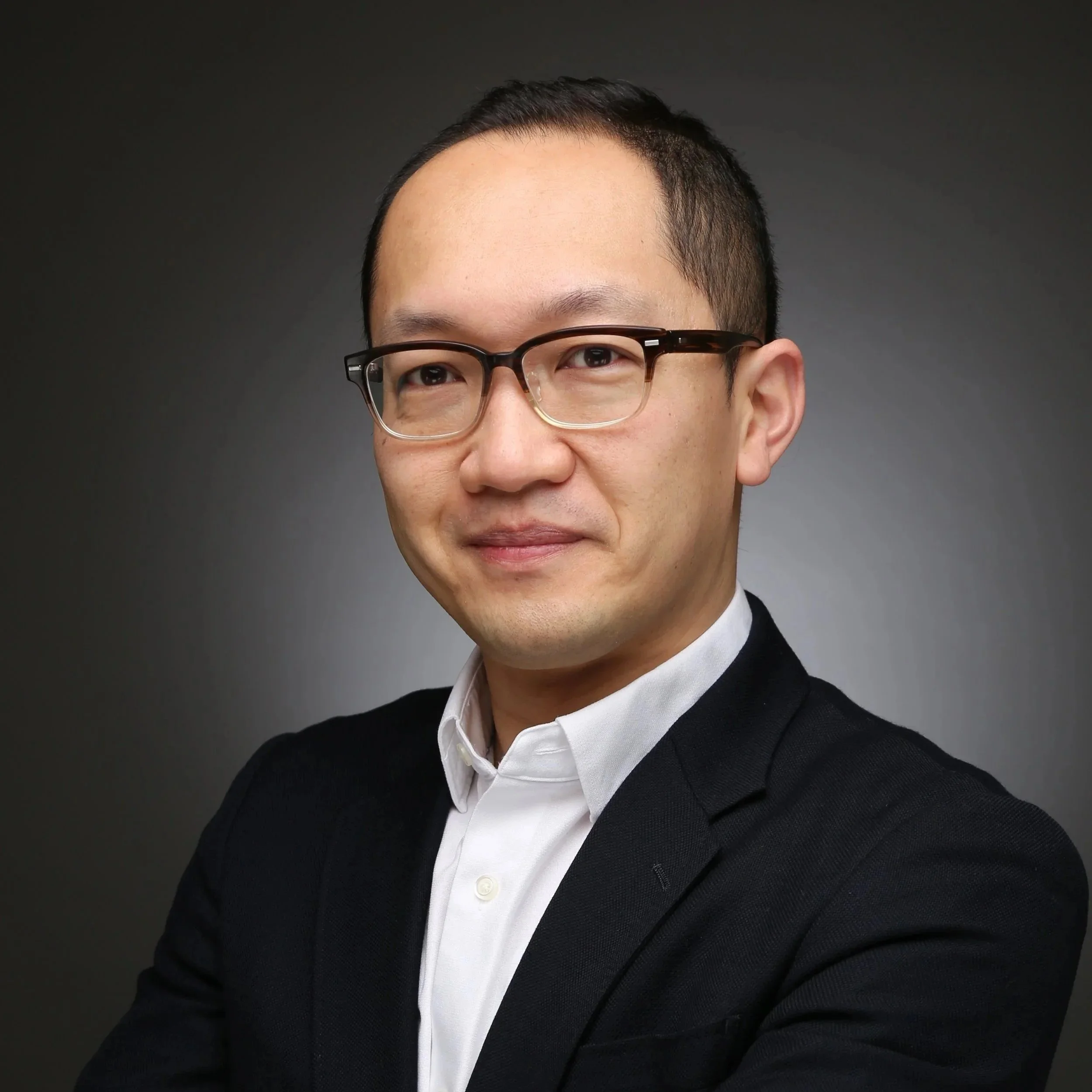 Close-up of a man with short dark hair, wearing glasses, a white shirt, and a black sweater against a dark background.