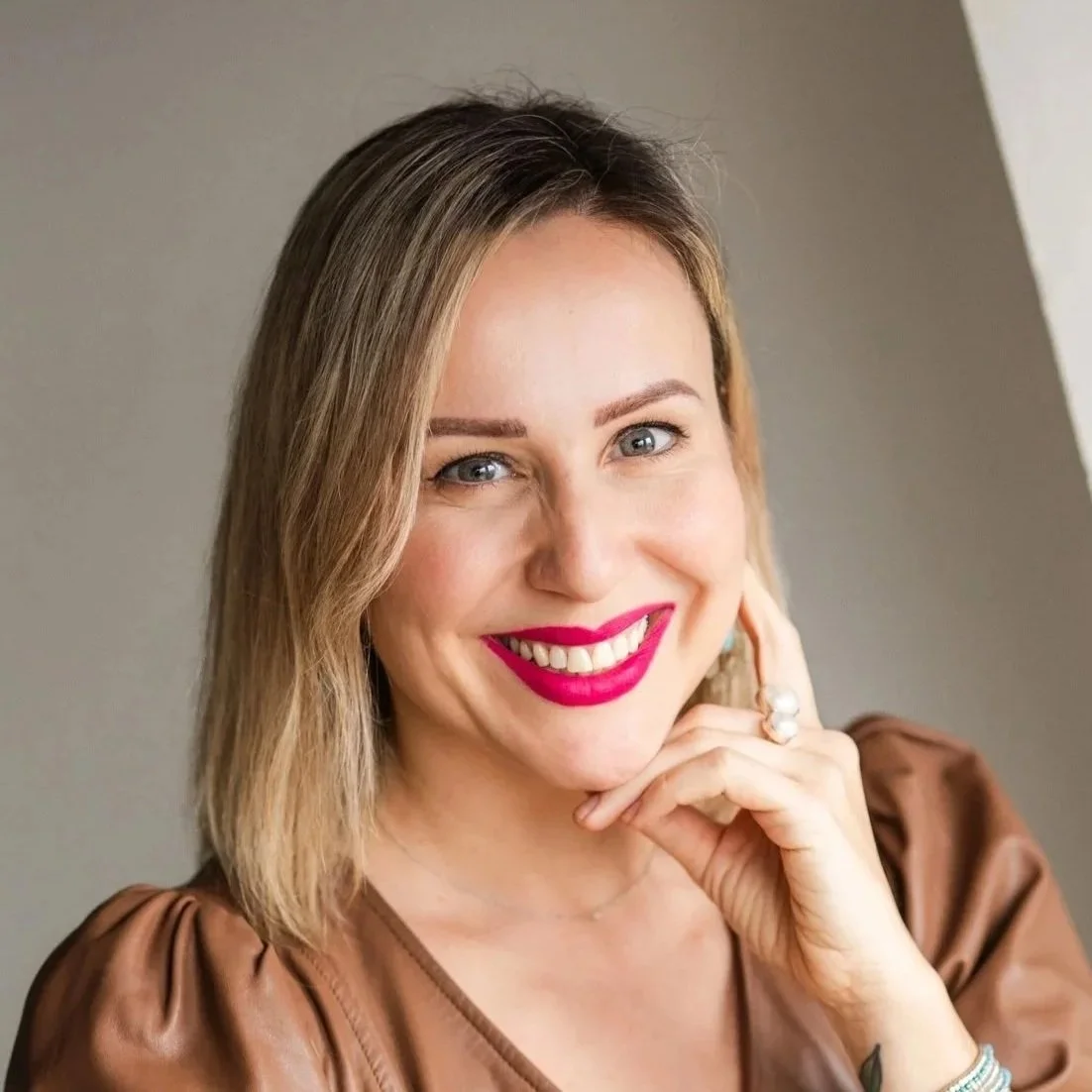 Close-up portrait of a woman with shoulder-length light brown hair, blue eyes, wearing bright pink lipstick, a brown top with puffed sleeves, and jewelry including rings and a bracelet, smiling and resting her chin on her hand.