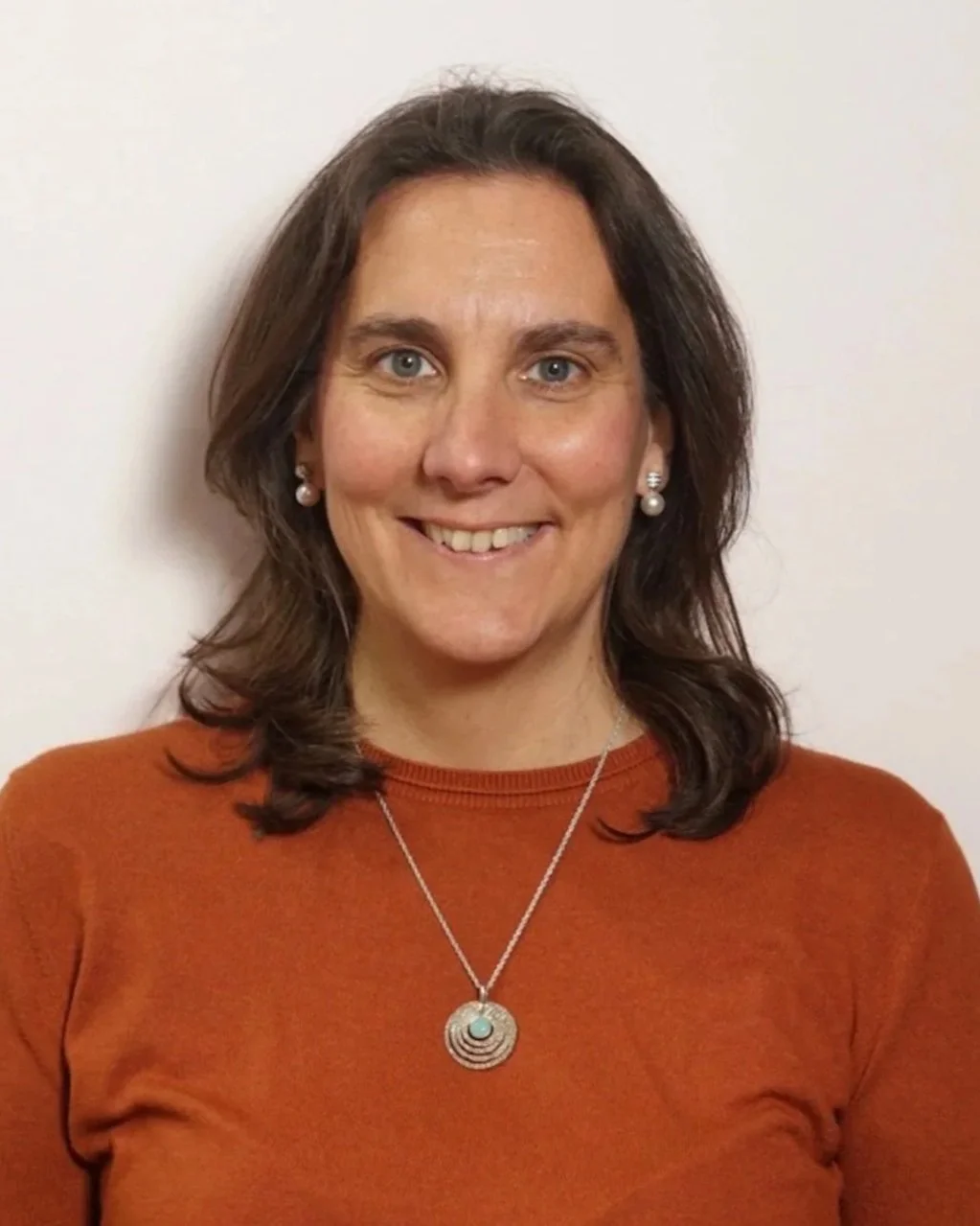 A woman with shoulder-length dark brown hair, blue eyes, and light skin is smiling and wearing pearl earrings, a silver necklace with a circular pendant, and an orange top. She is standing against a plain off-white wall.