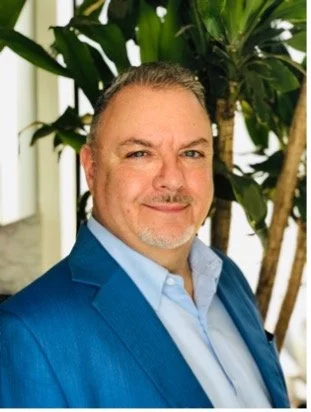 A middle-aged man with gray hair and a beard, wearing a blue blazer and light blue shirt, standing in front of large green plants indoors.