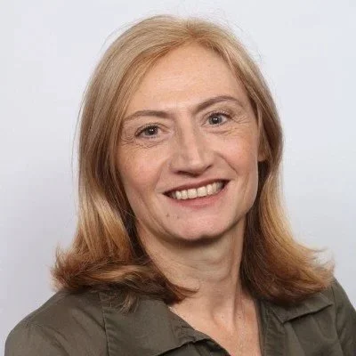 A smiling woman with shoulder-length blonde hair wearing a dark green shirt against a plain background.