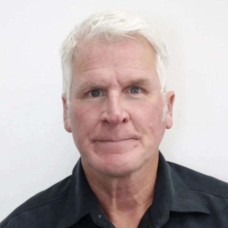 A middle-aged man with short, white hair and blue eyes, wearing a black collared shirt, standing against a plain white wall.