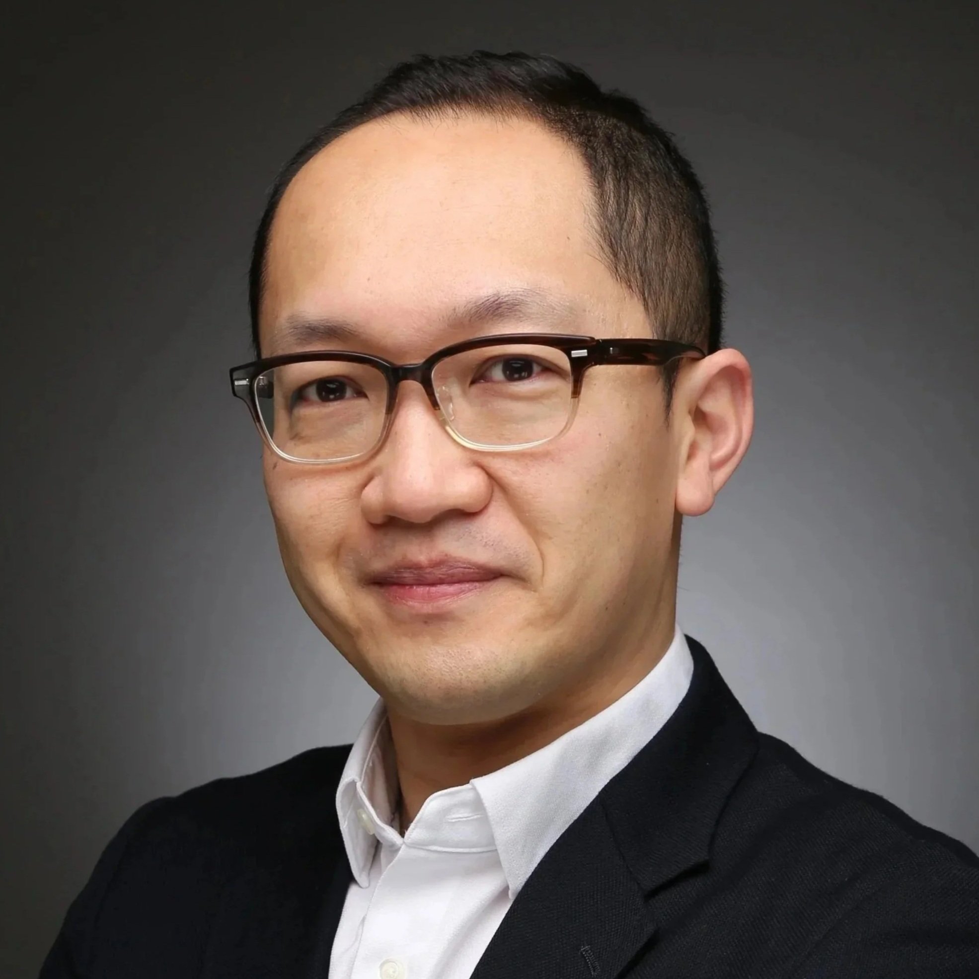 Close-up of a man wearing glasses, a white collared shirt, and a black blazer against a dark background.
