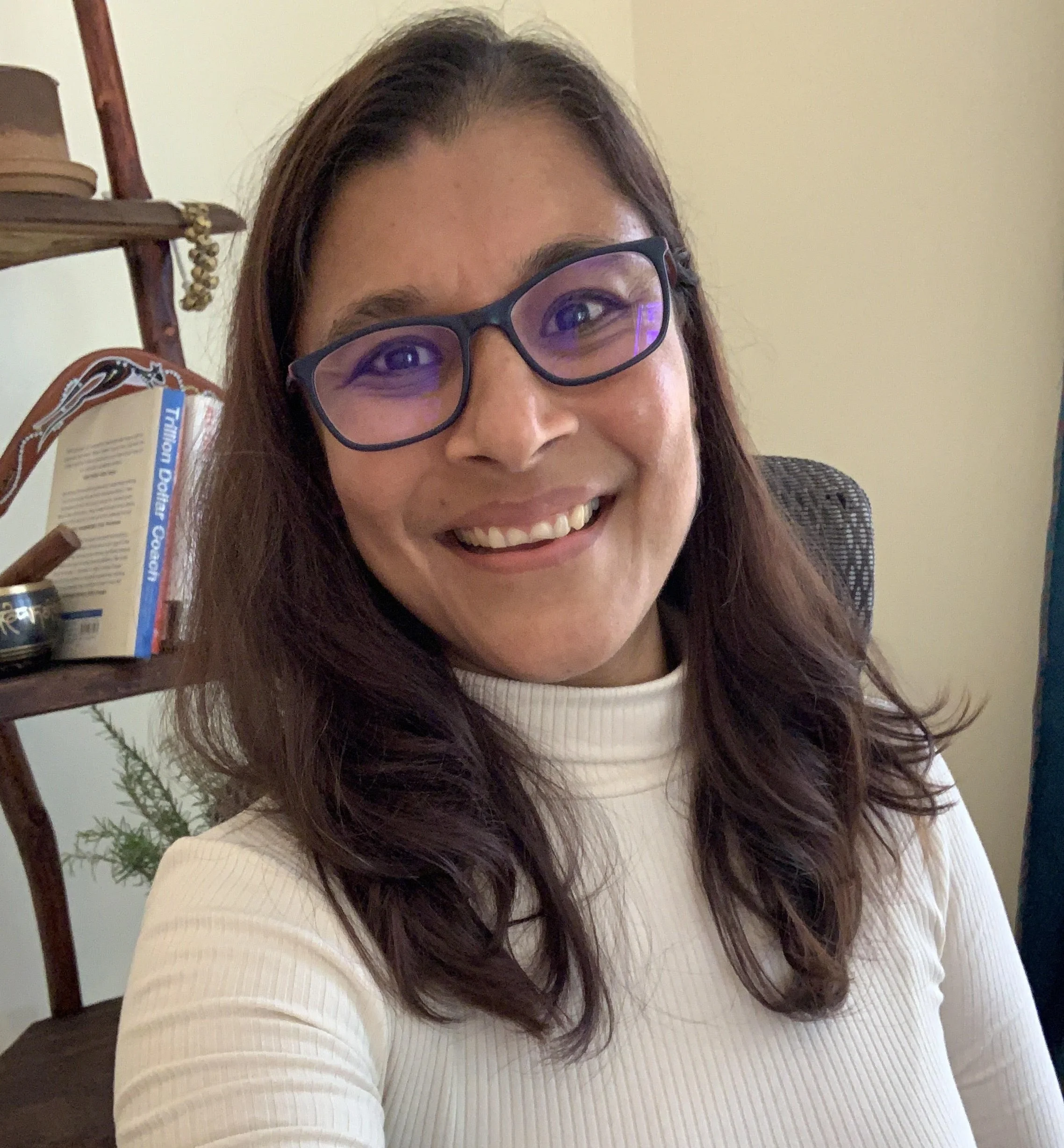 Close-up of a woman smiling, wearing glasses, with dark brown hair, in an indoor setting with a bookshelf and a plant in the background.